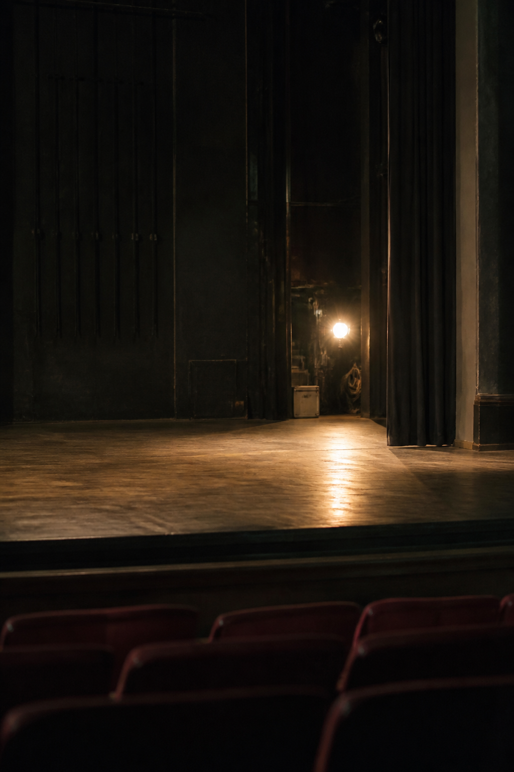 An empty theatre stage seen from the stalls, house lights half up and a bare wooden floor
