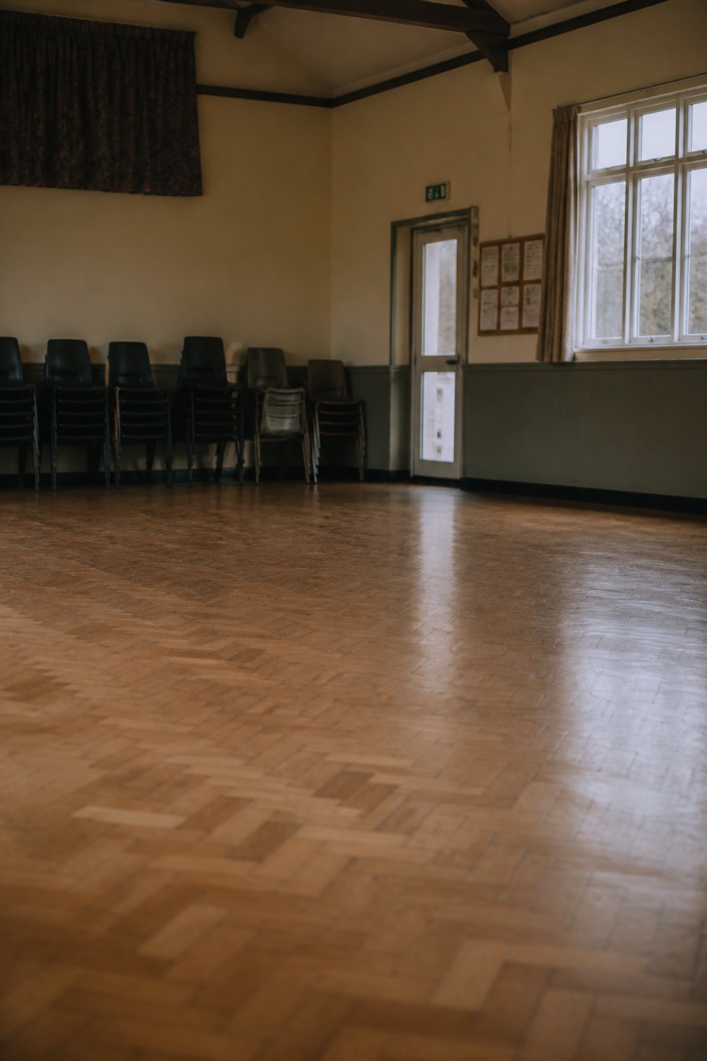 The empty interior of a village hall with a parquet floor, stacked chairs along one wall and high windows letting in grey daylight
