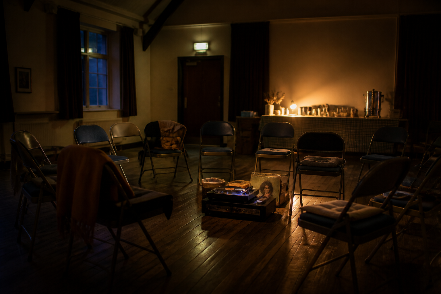 A small English village hall at night, chairs in a loose circle around a record player, warm lamps and a tea urn on a trestle table